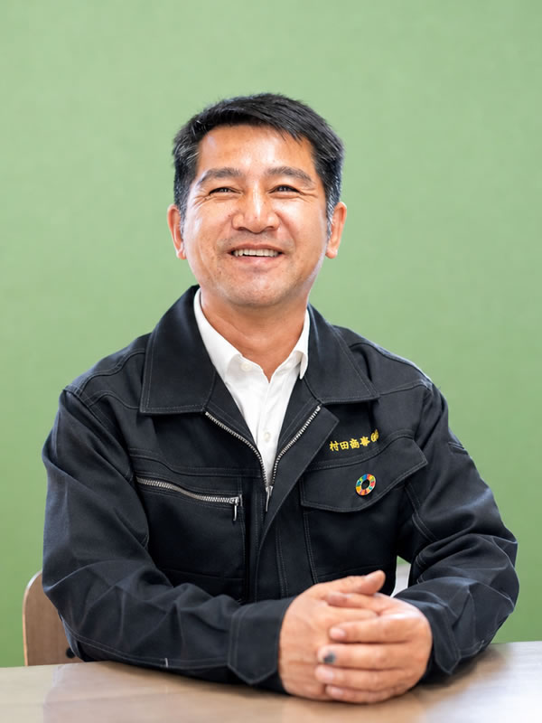 会社概要用の担当者ポートレート。黒い作業着を着た男性が机に手を置き微笑んでいる。